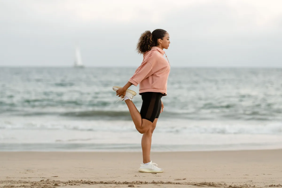A woman in a pink sweatshirt stands tall on a beach, holding one foot behind her in a quad stretch.