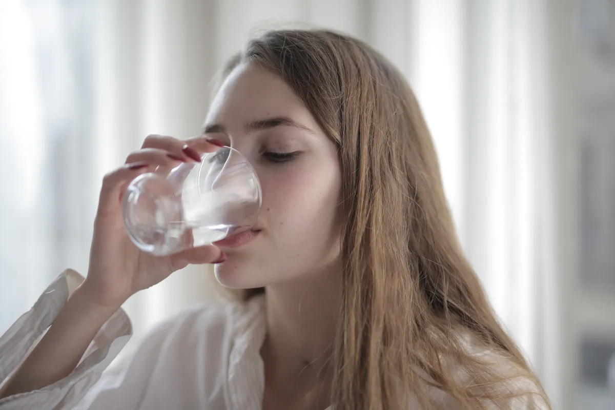 A woman sips a glass of water beside a bright window.