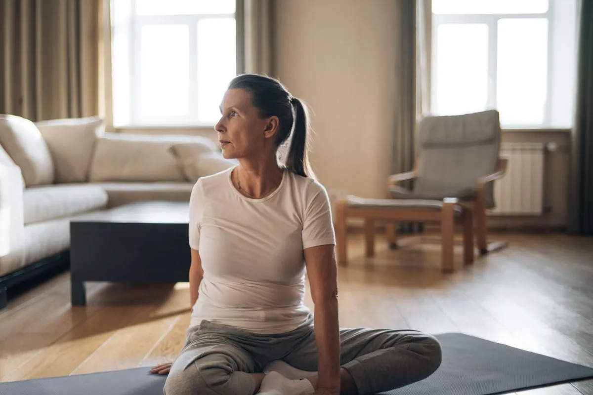 A woman sits cross-legged on an exercise mat in a sunlit living room.